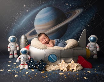 May include: A newborn baby sleeps in a spaceship-shaped bed, surrounded by astronaut figurines and star-shaped lights. The backdrop features a planet with rings and a starry sky, creating a space-themed nursery setting.
