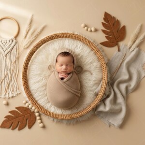 May include: A newborn baby wrapped in a beige swaddle and hat, nestled in a round woven basket on a fluffy white surface. The scene is decorated with dried plants, felt leaves, and a macrame wall hanging. The overall color palette is neutral.
