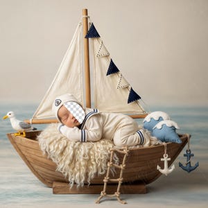 Op de afbeelding: Een slapende baby in een matrozenpak rust in een houten boot, compleet met een zeil en decoratieve elementen. De boot heeft een zeemeeuw, golfvormige kussens en een kleine touwladder. De scène roept een nautisch thema op.