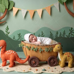 Op de afbeelding: Een pasgeboren baby slaapt in een houten wagen met een bruin en geel dinosaurus speelgoed en een oranje dinosaurus speelgoed. De wagen is versierd met groene bladeren en de achtergrond is een groene en bruine bos scène.