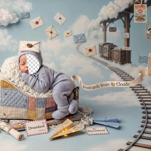 Op de afbeelding: Een pasgeboren baby in een blauwe outfit en muts, slapend in een patchworkmand. De scène heeft een spoorwegthema met papieren vliegtuigjes, enveloppen en de tekst "Postcards from Clouds" en "Dreamland".