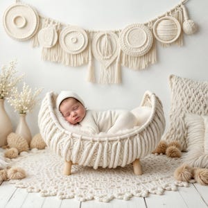 May include: A newborn baby sleeping in a white woven basket with a white crocheted blanket. The basket is on a white crocheted rug with white pom-poms around it. The background is a white wall with a white macrame wall hanging.