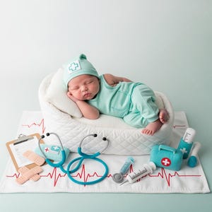 May include: A newborn baby dressed in a mint green medical outfit and hat, sleeping on a white quilted bed. Medical-themed props, including a stethoscope, bandages, and a toy medical kit, surround the baby on a white mat with a red heartbeat design.