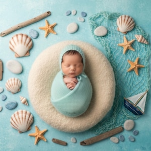 May include: A newborn baby wrapped in a light blue blanket and bonnet, resting on a cream-colored, circular cushion. The baby is surrounded by seashells, starfish, pebbles, driftwood, and a small sailboat on a light blue background.