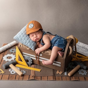 May include: A sleeping baby wearing a brown construction hat and denim overalls rests in a wooden toolbox. The toolbox is surrounded by miniature tools, blueprints, and a blue pillow, creating a construction theme.
