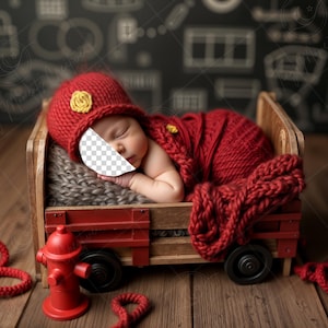 May include: A newborn baby in a red knitted hat with a yellow flower and a matching wrap, lying in a miniature wooden wagon. A red fire hydrant and rope are in the foreground. The wagon is on a wooden surface.