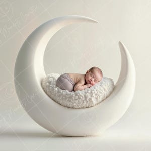 May include: A newborn baby sleeping on a white crescent moon shaped prop. The baby is wearing a light gray outfit and is curled up on a white fluffy cushion.