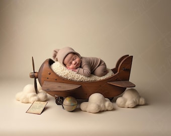 May include: A sleeping newborn in a beige outfit and hat rests in a wooden airplane prop. The plane has a propeller and wheels. White cloud props and a small globe are placed around the plane. A vintage-style card is also present.