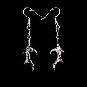 Puede incluir: Un par de pendientes de plata con diseño de alas. Los pendientes están sobre un fondo negro.