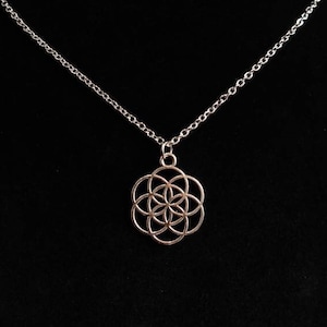 May include: A silver necklace with a pendant in the shape of a flower of life. The pendant hangs from a fine chain. The necklace is displayed against a black backdrop. The jewelry is a necklace.