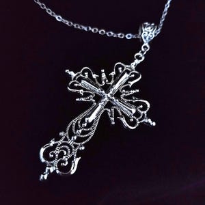 May include: Silver-toned filigree cross pendant necklace. The ornate cross features intricate openwork and a delicate chain. The pendant hangs from a fine chain, creating a gothic or vintage aesthetic.