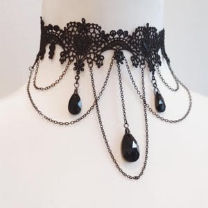 May include: A black lace choker necklace featuring an intricate floral pattern with scalloped edges. The design includes multiple thin black metal chains draped in layers, accented with three faceted black teardrop-shaped crystal pendants.