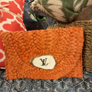 May include: Orange woven clutch bag with a white oval emblem featuring a black "LV" logo. The clutch is rectangular and appears to be made of a natural fibre. Other pillows and woven items are in the background.