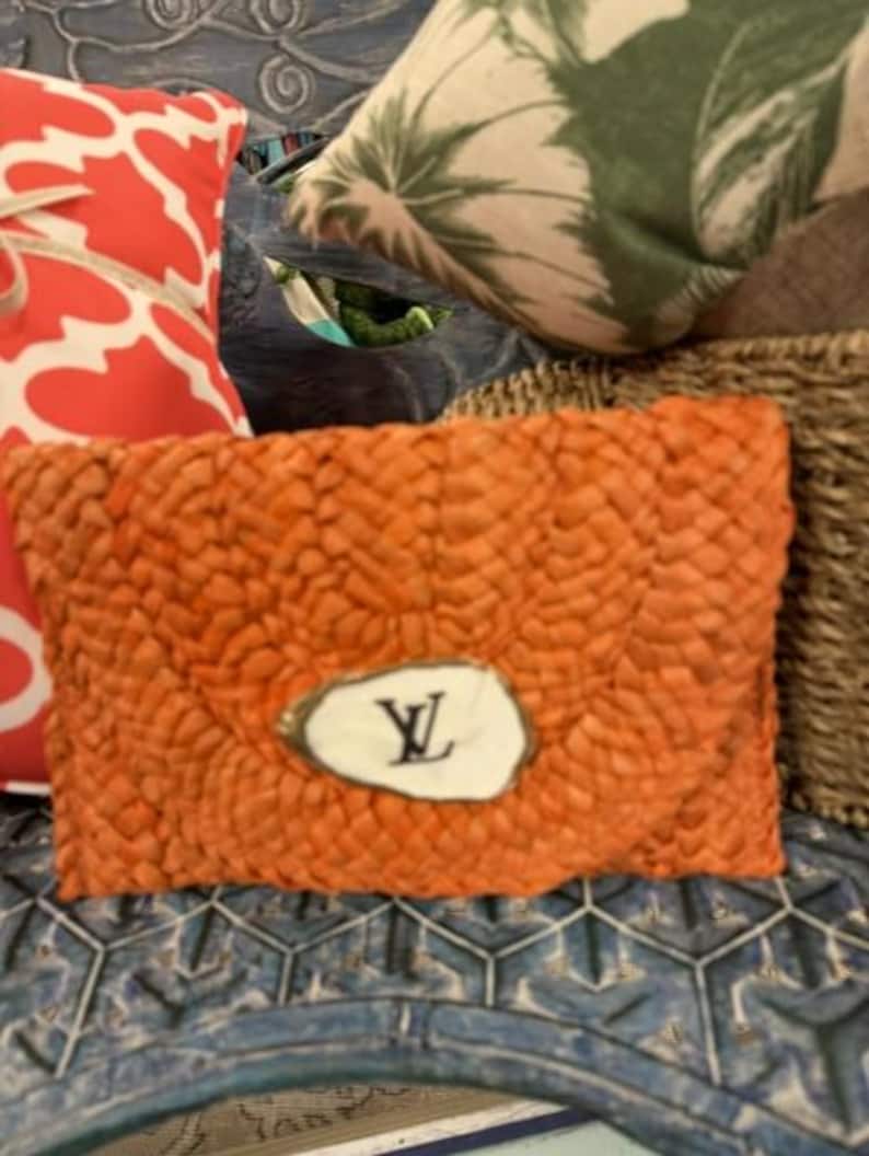 May include: Orange woven clutch bag with a white oval emblem featuring a black "LV" logo. The clutch is rectangular and appears to be made of a natural fibre. Other pillows and woven items are in the background.