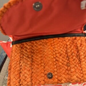 May include: Orange woven handbag with a red interior. The bag has a woven exterior, a magnetic snap closure, and a black trim. The interior is a solid red colour. The bag is open, revealing the interior.
