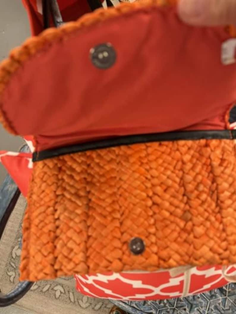 May include: Orange woven handbag with a red interior. The bag has a woven exterior, a magnetic snap closure, and a black trim. The interior is a solid red colour. The bag is open, revealing the interior.