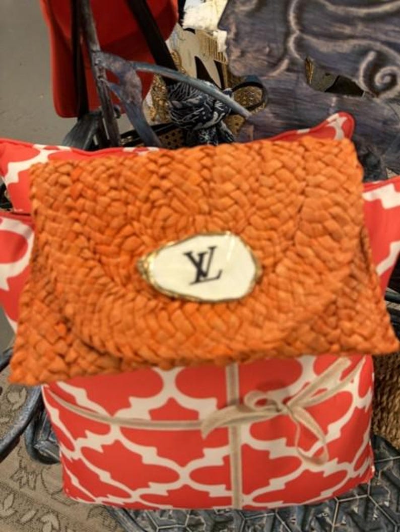 May include: An orange woven clutch bag with a white and gold emblem featuring black lettering. The clutch rests on a red and white patterned cushion, tied with a beige ribbon. The theme is accessories.