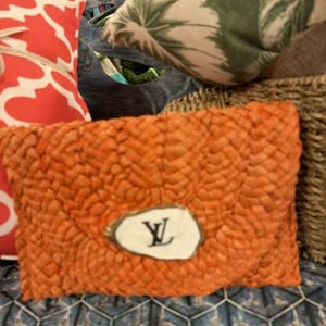 May include: Orange woven clutch bag with a white oval emblem featuring a black "Y" design. The clutch is rectangular and has a textured surface. Other items in the background include a red and white patterned cushion and a cushion with a green leaf design.