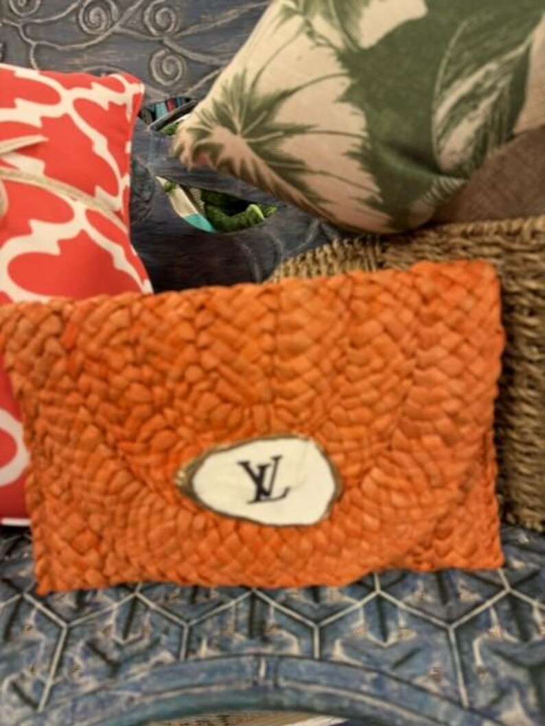 May include: Orange woven clutch bag with a white oval emblem featuring a black "Y" design. The clutch is rectangular and has a textured surface. Other items in the background include a red and white patterned cushion and a cushion with a green leaf design.