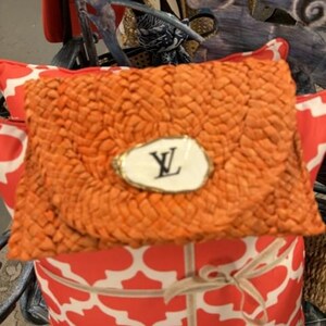 May include: An orange woven clutch bag with a white and gold emblem featuring black lettering. The clutch rests on a red and white patterned cushion, tied with a beige ribbon. The theme is accessories.