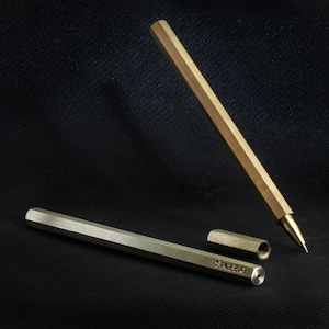 May include: A brass pen with a hexagonal barrel and a silver cap. The pen is lying on a black surface, and the cap is off. The pen is engraved with the word "STEEBR".