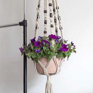 May include: A beige macrame plant hanger with a terracotta pot filled with vibrant purple petunias. The hanger features wooden beads and a tassel, adding a bohemian touch. Perfect for indoor or outdoor decor.