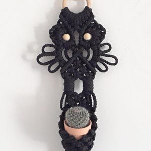 May include: Black macrame plant hanger with a wooden ring and two wooden beads. The hanger holds a small terracotta pot with a cactus. The bottom of the hanger has a tassel. The hanger is hanging on a white wall.