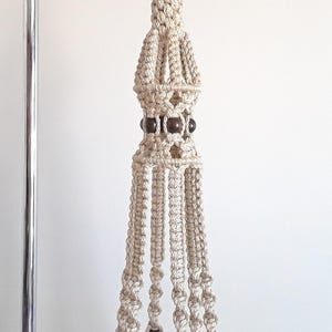 May include: A beige macrame plant hanger with dark brown wooden beads. The handmade plant holder is suspended from a metal hook and features a woven design with multiple strands. This decorative item is perfect for displaying plants.