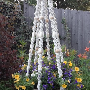 May include: A cream-colored macrame plant hanger featuring twisted cord patterns and smooth, round wooden beads integrated into the vertical strands. The hanger supports a round pot filled with yellow and purple flowers.