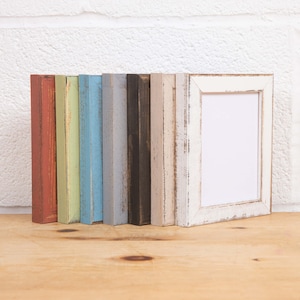 May include: A set of eight picture frames in various colours, including red, green, blue, grey, black, and white. The frames are all made of wood and have a distressed finish. The frames are arranged in a row on a wooden surface.
