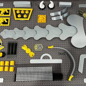 Op de afbeelding: Een verzameling 3D-geprinte onderdelen in grijs, geel en zwart. De componenten, waaronder gebogen en geometrische vormen, zijn gerangschikt op een patroon. De onderdelen suggereren een groter bouwproject met gedetailleerde elementen.