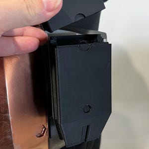 Puede incluir: Una caja de plástico negra con una tapa con bisagras, unida a un objeto marrón y negro. La caja está abierta y la tapa está siendo sostenida por una mano.