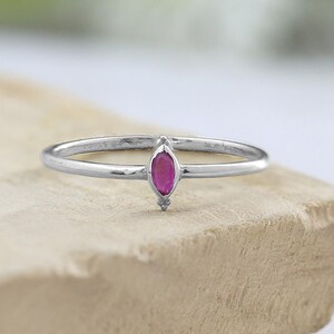 May include: A silver ring with a marquise-cut, deep pink gemstone. The ring is set on a light-colored, textured surface, with a blurred green and white background. The gemstone is the focal point of the delicate, minimalist design.