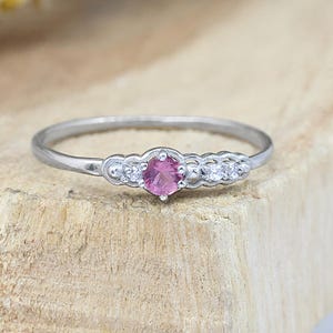 Puede incluir: Un anillo plateado con una piedra rosa central y pequeñas piedras blancas. El anillo está sobre una superficie de madera, con un elemento floral amarillo borroso en el fondo. El diseño del anillo es delicado y elegante.