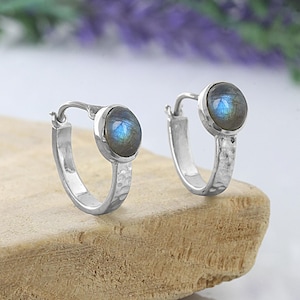 May include: A pair of silver hoop earrings featuring a hammered texture on the band and a round, cabochon-cut labradorite gemstone set at the top. The stones display a distinct blue iridescence, and the hoops have a secure hinged closure.