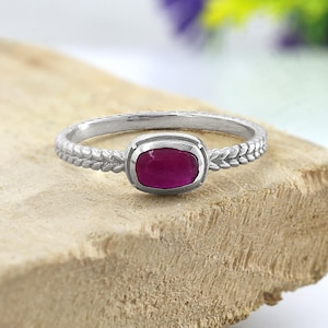 Puede incluir: Un anillo de plata con una gema rectangular de color rojo intenso. La banda presenta un delicado patrón de hojas a cada lado de la gema. El anillo se muestra sobre una superficie texturizada de color marrón claro, con vegetación borrosa en el fondo.