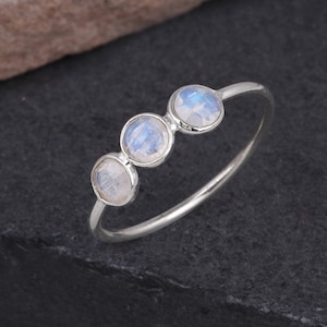 Könnte beinhalten: Ein Silberring mit drei runden, facettierten Mondstein-Edelsteinen. Die Edelsteine haben eine hellblaue und weiße Farbe und sind in einem schlichten, zarten Band gefasst. Der Ring wird auf einer dunklen, strukturierten Oberfläche präsentiert.