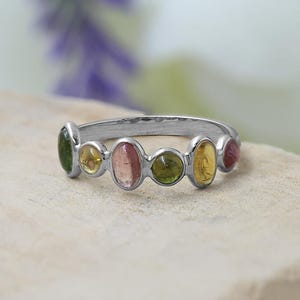 Peut inclure: Une bague en argent ornée d'une rangée de pierres précieuses colorées. Les pierres, de forme ovale, présentent des teintes vertes, jaunes, roses et rouges. La bague est posée sur une surface claire, avec un fond floral flou.