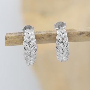 May include: A pair of silver hoop earrings featuring a textured, repeating leaf or wheat pattern along the outer surface. The earrings are circular in shape with a polished metallic finish, showcasing a detailed, embossed design that wraps around the hoop.