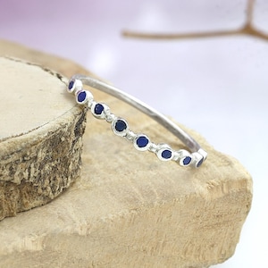 Op de afbeelding: Een zilveren ring met een fijne band, versierd met een reeks kleine, ronde, diepblauwe edelstenen. De ring wordt gepresenteerd op een lichtbruin, getextureerd oppervlak, dat het elegante ontwerp en vakmanschap benadrukt.