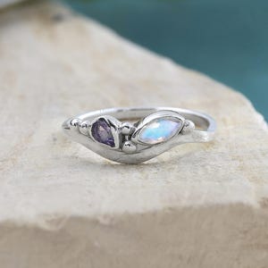 May include: A silver ring featuring a purple gemstone and a pearlescent, oval-shaped stone. The ring has a curved design with small, round accents. The ring is displayed on a light-colored, textured surface.