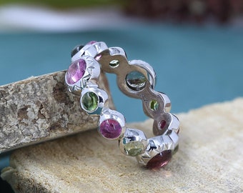 May include: A silver ring with a repeating pattern of colorful gemstones. The ring features alternating pink, green, and other colored stones set in a polished silver band. The gemstones are round and evenly spaced around the ring.