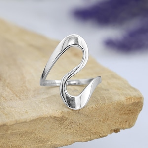 Pode incluir: Um anel prateado com um design fluido e abstrato. O anel apresenta uma forma contínua, semelhante a uma onda, que se curva e se dobra, criando uma estética aberta e moderna. O anel é exibido em uma superfície de madeira marrom clara.