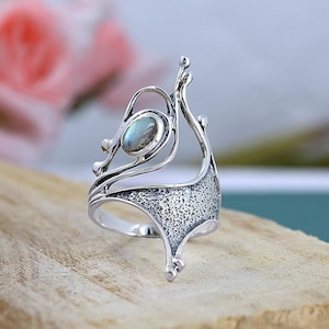 Peut inclure: Une bague en argent avec une pierre précieuse ovale et irisée. La bague présente un design ajouré orné avec des détails texturés et une forme artistique unique. La pierre est sertie dans un sertissage à lunette, et la bague a une finition polie.