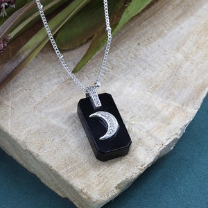May include: A silver necklace with a rectangular black pendant. The pendant features a crescent moon design with embedded clear stones. The necklace is displayed on a light-colored wooden surface.