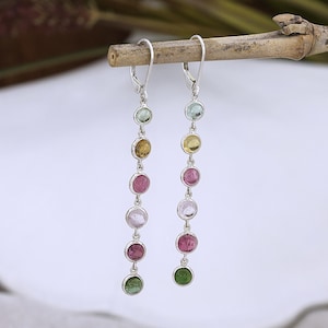May include: Dangle earrings with a silver setting, each featuring a series of colourful, round gemstones. The gemstones are in shades of green, yellow, pink, and clear. The earrings are suspended from a lever-back closure.