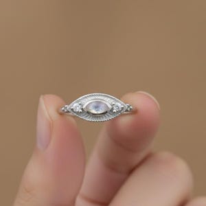 以下が含まれることがあります： 中央に楕円形のムーンストーン、両側に2つの小さなクリアストーンをあしらったシルバーリング。 リングは詳細なテクスチャード加工のバンドです。 背景はニュートラルです。