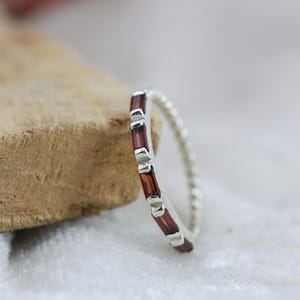 May include: A silver ring with a beaded band. The ring is adorned with a line of rectangular, dark red gemstones set in silver. The ring is presented on a light-coloured surface, highlighting its design and colour.