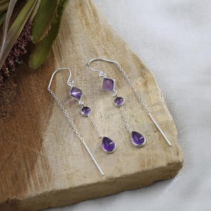 Puede incluir: Un par de pendientes colgantes de plata con piedras de amatista. Cada pendiente presenta una piedra púrpura en forma de diamante, redonda y de lágrima. Los pendientes se exhiben sobre una superficie de madera.