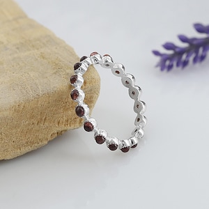Op de afbeelding: Een zilveren ring met een doorlopende rij kleine, ronde, donkerrode edelstenen. De ring staat tegen een lichte achtergrond met een stuk bruine steen en een takje paarse bloemen.